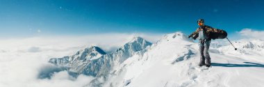Snowboardcu karlı zirvede, panoramik manzarada duruyor. Parlak mavi gökyüzü ve beyaz kar hakim. Kış sporları veya seyahat için ideal. Kopyalama alanı olan geniş pankart.
