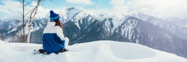 Kış giysileri içindeki bir kadın huzur içinde oturup karlı bir dağ manzarasına hayran kalır. Zengin maviler ve beyazlar baskın. Seyahat ve tatil temaları için mükemmel. Geniş pankart.