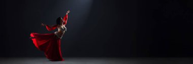 Karanlık arka planda canlı kırmızı elbisesi olan zarif bir dansçı. Zarif poz ve akıcı kumaş çarpıcı bir kontrast yaratır, sanatsal tasarımlar için idealdir. Kopyalama alanı olan geniş pankart.