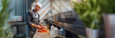 Şef modern bir mutfakta pizza yapıyor, afiş geniş. Sıcak renkler ve profesyonel kurulum, aşçılık sektörü promosyonları için uygun.