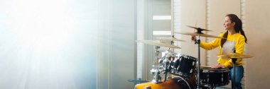 Parlak bir stüdyoda davul çalan genç bir kadın. Sıcak renkler ve modern tarz, müzik temalı afişler ve reklamlar için uygun. Kopyalama alanı olan geniş pankart.