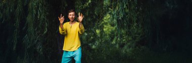 Sarı gömlekli bir adam yapraklı dalların altında kendini ifade eder. Canlı renkler, doğal ayar, metinli reklamlar için uygun. Doğa ve refah temaları için ideal.