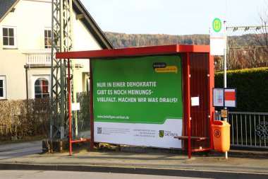 Aue-Bad Schlema, Germany - March 26, 2024: Poster on a bus stop, promoting democracy and inviting citizens to participate in social programs led by the government of Saxon clipart