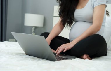 Yatakta dizüstü bilgisayar kullanan hamile bir kadın.
