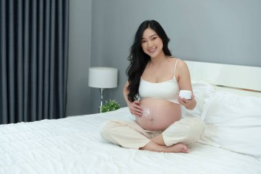 Hamile bir kadın karnına krem sürüyor. Güzellik için. Yatakta nemlendirici bir cilt.