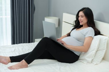 Yatakta dizüstü bilgisayar kullanan hamile bir kadın.