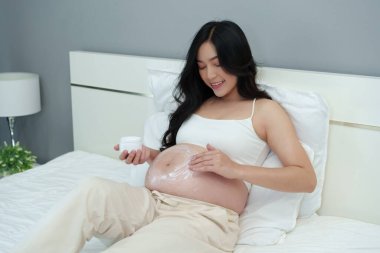 Hamile bir kadın karnına krem sürüyor. Güzellik için. Yatakta nemlendirici bir cilt.