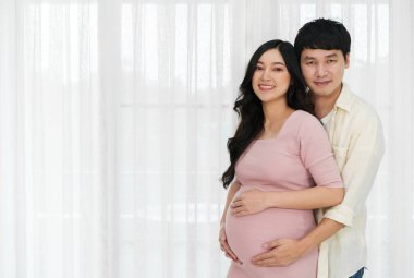 Evli çift bebek bekliyor. Erkek, hamile karısını pencere arkasında kucaklıyor.