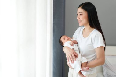 mother holding her newborn baby and looking through the window in bedroom