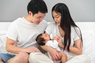 parents (father and mother) feeding milk bottle to newborn baby on a bed