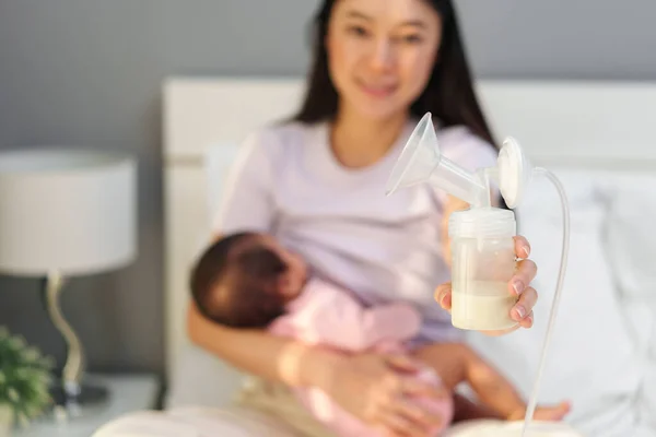 mother holding breast pump milk in the bottle while her breastfeeding newborm baby