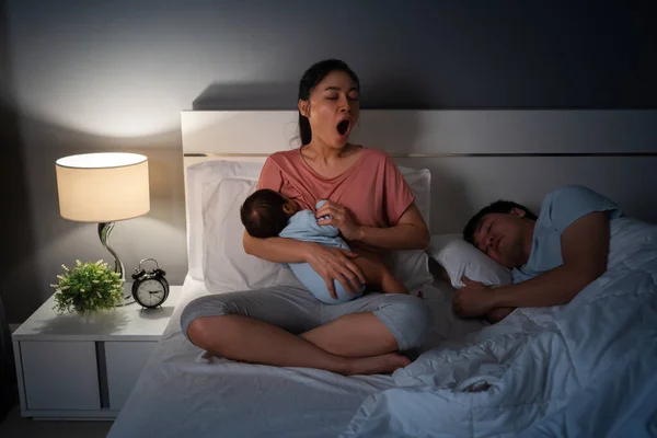 tired mother yawning and breastfeeding newborm baby while her husband sleeping on a bed at night