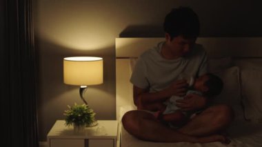 Stresli baba, ağlayan bebeğini gece yatağında süt şişesiyle beslemeye çalışıyor.