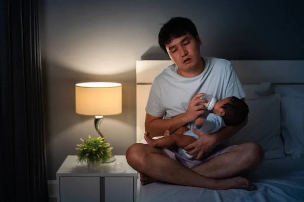 sleepy and tired father feeding milk bottle to newborn baby on a bed at night