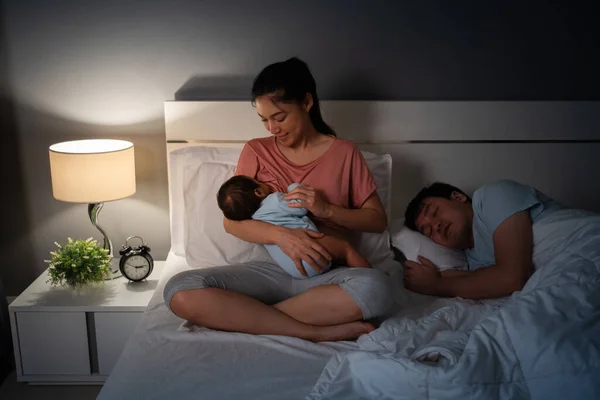 stock image mother breastfeeding newborm baby while her husband sleeping on a bed at night