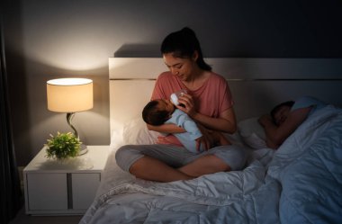 Anne, yeni doğan bebeğe süt içirirken kocası geceleri yatakta uyuyor.