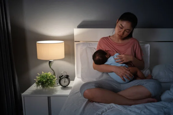 sleepy and tired mother breastfeeding newborm baby on a bed at night