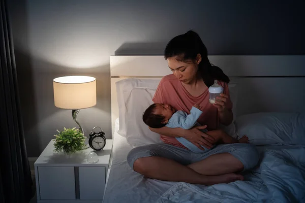 stressed mother trying to feeding milk bottle to her crying newborn baby on a bed at night