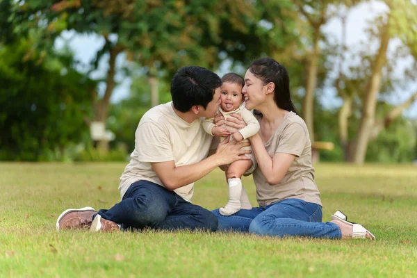 happy family sitting on a green grass filed. father and mother kissing their infand baby in the park