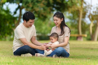 Parkta çimlerin üzerinde oturan mutlu bir aile. Baba ve anne bebek bebekle konuşuyor ve oynuyor..