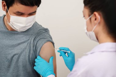 Doktor elinde şırınga tutuyor ve hasta adama tıbbi maske takıyor. Covid-19 veya koronavirüs aşısı