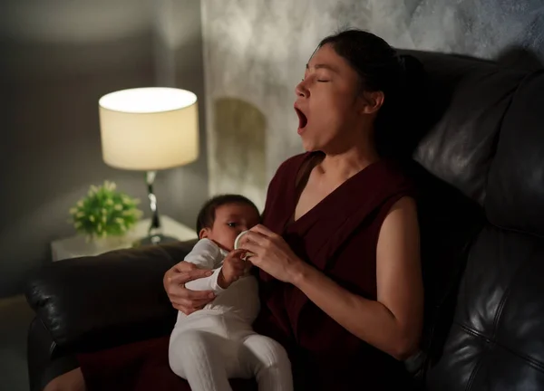 sleepy mother yawning and feeding milk bottle to infant baby on a sofa in the living room at night