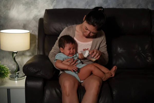 stressed mother trying to feeding milk bottle to crying infant baby on a sofa in the living room at night