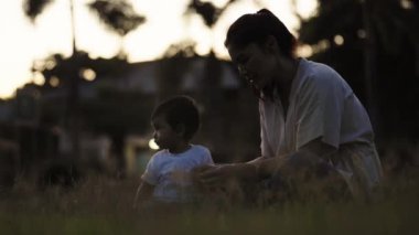 Anne ve bebek gün batımında parkta çimlerin üzerinde oturup oynuyorlar.