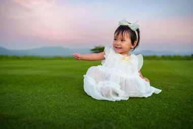 Beyaz elbiseli mutlu bir kız bebek gökyüzü ve dağ arkası olan bir çimenlikte oturuyor.