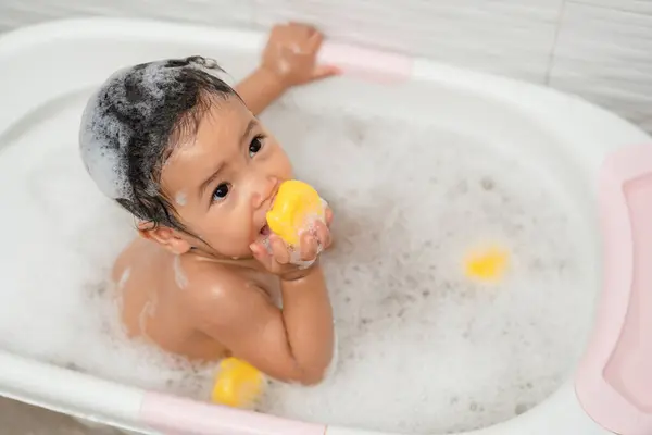 happy infant baby take a bath and playing rubber duck with foam bubbles in bathtub at home