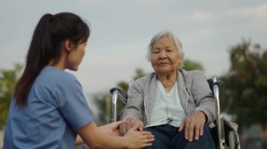 Hemşire, parkta tekerlekli sandalyedeki hasta dost canlısı bir bakıcı iken yaşlı bayanla diz ağrısı hakkında konuşup ilgilenir.