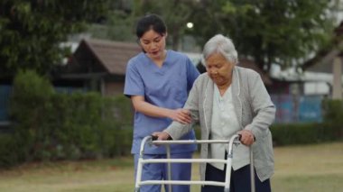Bakıcı hemşire, parkta Walker 'la yürüyen yaşlı kadına destek oluyor.