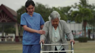 Bakıcı hemşire, parkta Walker 'la yürüyen yaşlı kadına destek oluyor.