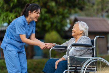 Mutlu hemşire, parkta tekerlekli sandalyedeki yaşlı kadın hastayla ilgileniyor ve onunla konuşuyor.