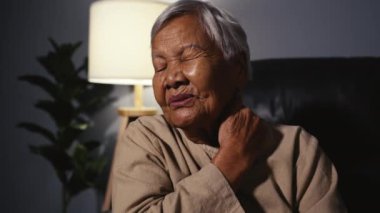 Geceleyin oturma odasında kanepede otururken boynu ağrıyan yaşlı bir kadın.