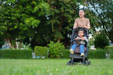 Mutlu anne yeni yürümeye başlayan bebek arabasını itiyor ve parkta yürüyor.