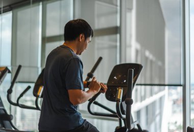 Genç adam eliptik bir çapraz antrenör üzerinde egzersiz yapıyor ve spor salonunda performans için akıllı saati kontrol ediyor.