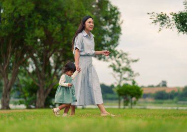 Mutlu anne küçük kızıyla parkta çim tarlasında yürüyor.