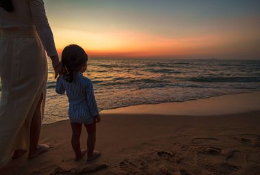 Anne, Pattaya, Tayland 'da gün batımında küçük bebekle birlikte deniz sahilinde duruyor.