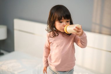 Küçük kız yatakta şişeden süt içiyor.