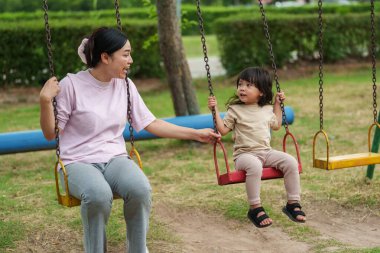 Neşeli küçük kız ve annesi açık hava çocuk parkında salıncakta oynuyor.