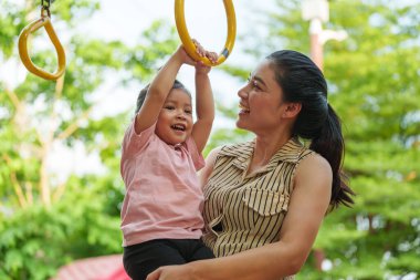 Anne kızını oyun sahasındaki yüzüklere asmak için tutuyor.
