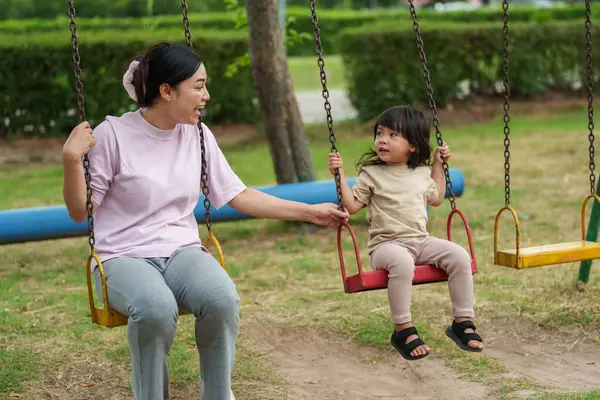 Neşeli küçük kız ve annesi açık hava çocuk parkında salıncakta oynuyor.