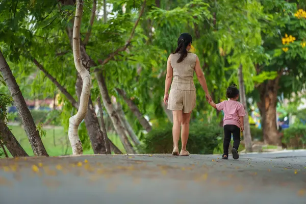 Yürüyen ve parktaki kaldırımda annesiyle el ele tutuşan küçük bir kız.