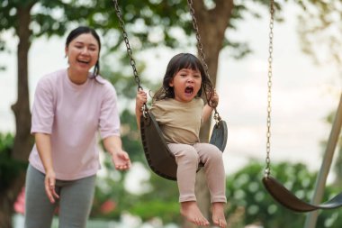 Neşeli küçük kız ve annesi açık hava çocuk parkında salıncakta oynuyor.