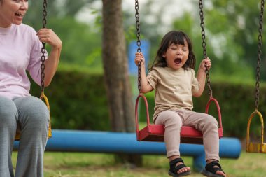 Neşeli küçük kız ve annesi açık hava çocuk parkında salıncakta oynuyor.