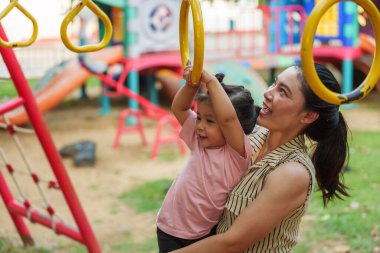 Anne kızını oyun sahasındaki yüzüklere asmak için tutuyor.