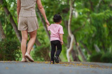 Yürüyen ve parktaki kaldırımda annesiyle el ele tutuşan küçük bir kız.