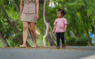 Küçük kız annesiyle parkta kaldırımda yürüyor.