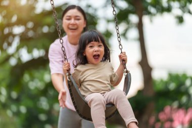 Neşeli küçük kız ve annesi açık hava çocuk parkında salıncakta oynuyor.